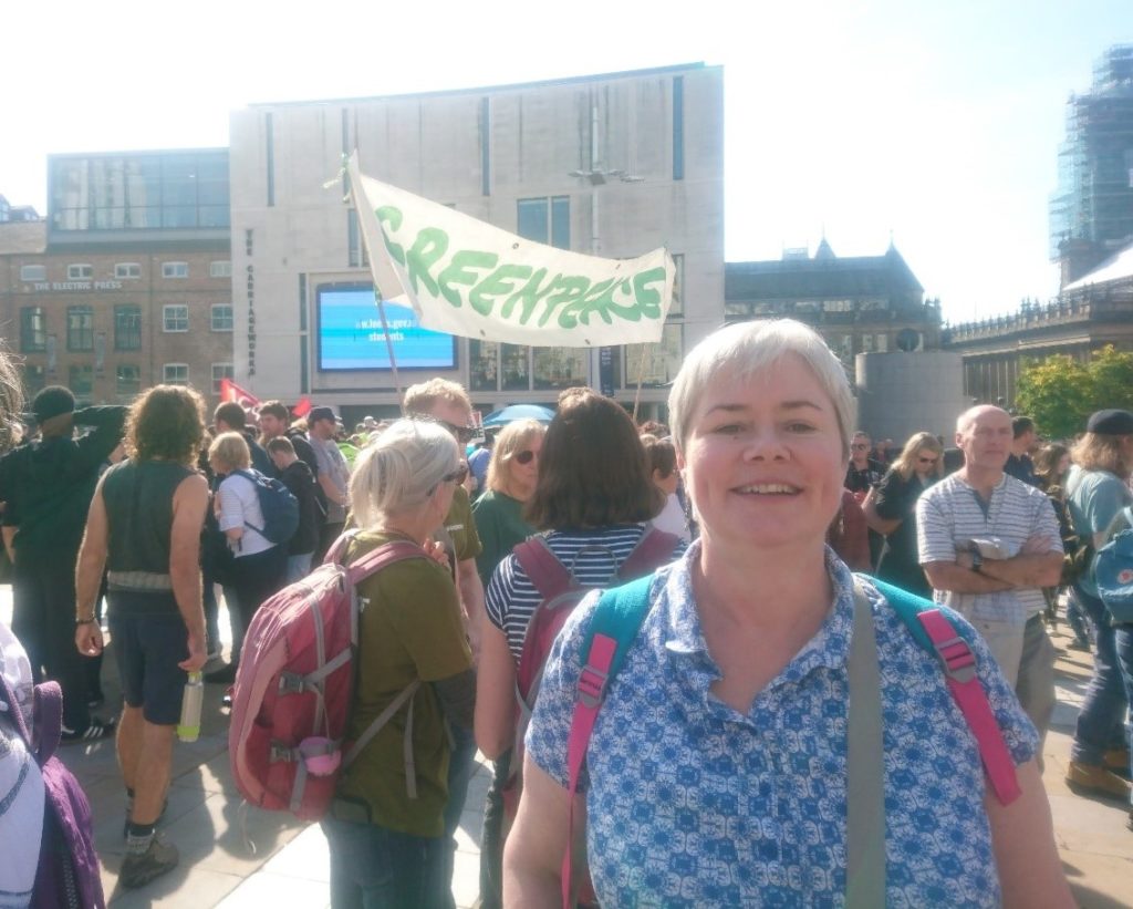 a woman stood in Millenium Square with a crowd of people and a large Greenpeace banner in the background
